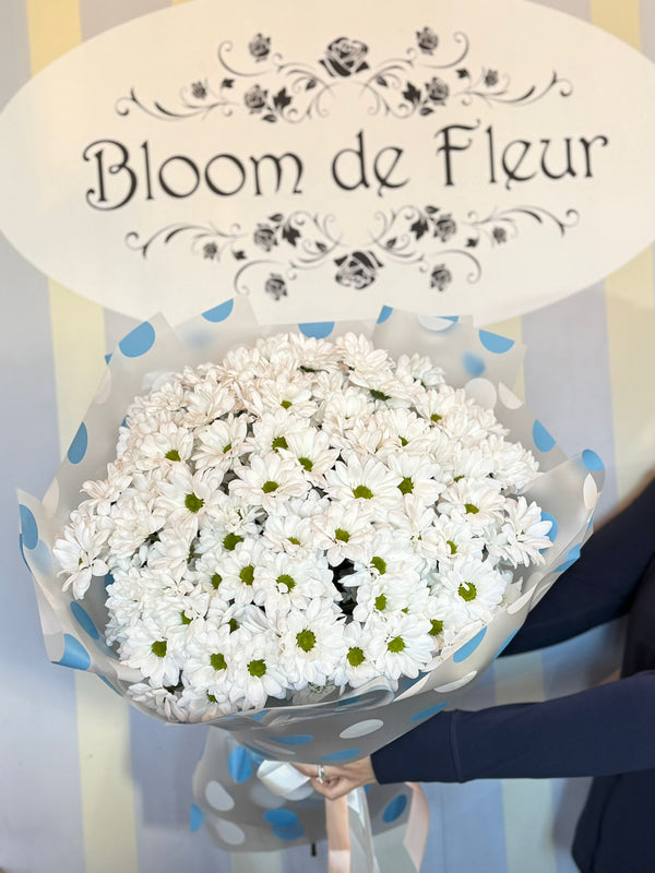 Bouquet of white daisy chrysanthemums with green centers wrapped in gray and blue polka-dot paper, tied with ribbon.