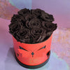 A round red Bloom de Fleur gift box with butterfly‑shaped cutouts and a black base is filled with large deep black preserved roses and sits on a lavender‑toned textured surface against a pastel purple backdrop.