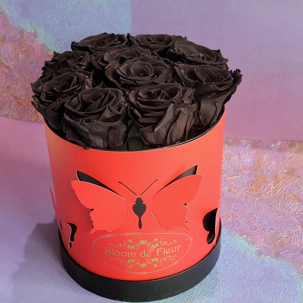 A round red Bloom de Fleur gift box with butterfly‑shaped cutouts and a black base is filled with large deep black preserved roses and sits on a lavender‑toned textured surface against a pastel purple backdrop.