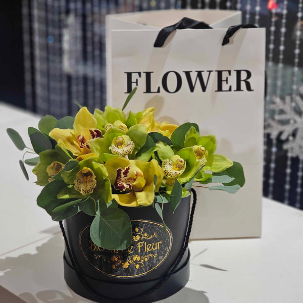 A matte black round gift box bearing a gold “Bloom de Fleur” logo holds a compact arrangement of chartreuse‑green cymbidium orchids and silvery eucalyptus leaves; it sits on a white tabletop in front of a white “FLOWER” paper bag and a backdrop of beaded curtains and hanging silver snowflake decorations.