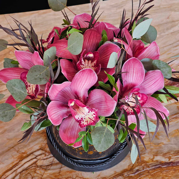 A matte black round gift box with a gold “Bloom de Fleur” logo and black rope handles sits on a wood‐grain tabletop, overflowing with large mauve‑pink cymbidium orchids, silvery eucalyptus leaves, and slender deep burgundy foliage; in the background, a black chair and a pale wall striped in cream and gray add subtle contrast.