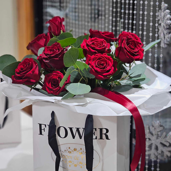A white square paper gift bag with black ribbon handles and the word “FLOWER” printed in bold black letters holds a lush bouquet of deep red roses and silvery-green eucalyptus leaves, all wrapped in crisp white tissue paper and tied with a burgundy satin ribbon; in the softly blurred background, a second matching bag sits beside a window draped with beaded curtains and hanging silver snowflake ornaments.