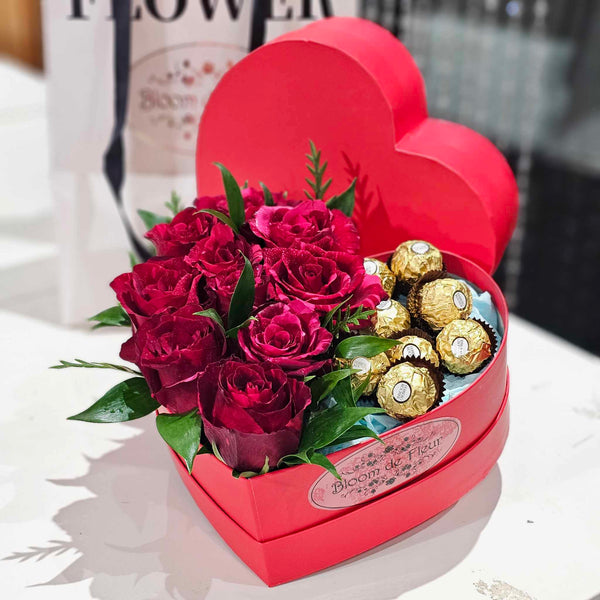 Heart‑shaped red Bloom de Fleur gift box overflowing with deep red roses, lush green foliage, and gold‑wrapped Ferrero Rocher chocolates nestled on blue tissue—an elegant Valentine’s floral and chocolate arrangement