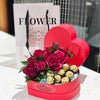 Heart‑shaped red Bloom de Fleur gift box overflowing with deep red roses, lush green foliage, and gold‑wrapped Ferrero Rocher chocolates nestled on blue tissue—an elegant Valentine’s floral and chocolate arrangement