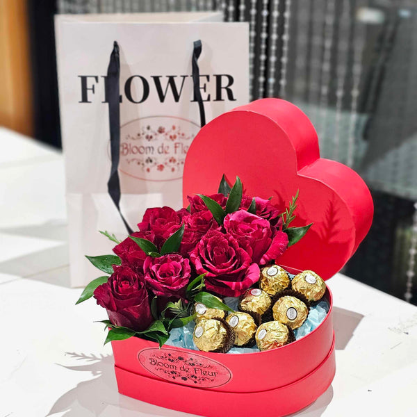 Heart‑shaped red Bloom de Fleur gift box overflowing with deep red roses, lush green foliage, and gold‑wrapped Ferrero Rocher chocolates nestled on blue tissue—an elegant Valentine’s floral and chocolate arrangement