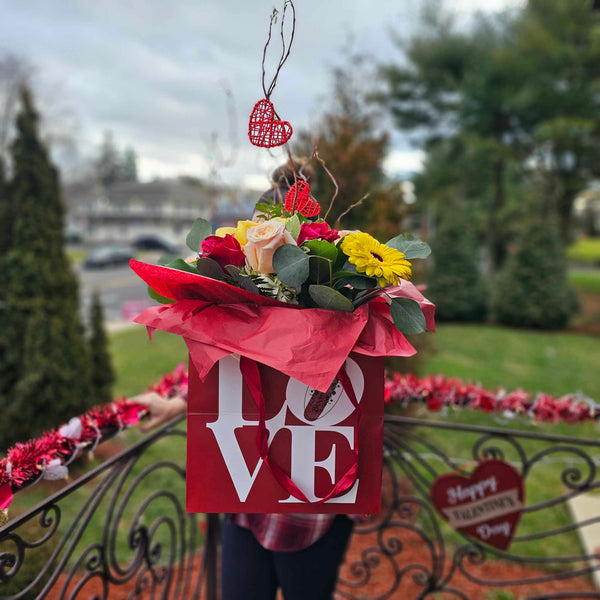 A red square gift box printed with the word “LOVE” in white holds a vibrant bouquet of red and pale pink roses, yellow gerbera daisies, yellow orchids and assorted greenery, all wrapped in red tissue paper and accented with red wicker heart decorations