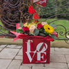 A red square gift box printed with the word “LOVE” in white holds a vibrant bouquet of red and pale pink roses, yellow gerbera daisies, yellow orchids and assorted greenery, all wrapped in red tissue paper and accented with red wicker heart decorations, set on a stone‐tiled patio.