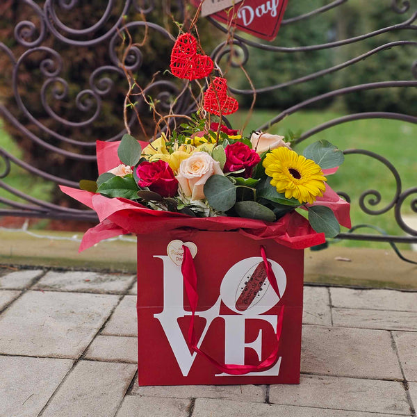 A red square gift box printed with the word “LOVE” in white holds a vibrant bouquet of red and pale pink roses, yellow gerbera daisies, yellow orchids and assorted greenery, all wrapped in red tissue paper and accented with red wicker heart decorations, set on a stone‐tiled patio.