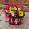 A red square gift box printed with the word “LOVE” in white holds a vibrant bouquet of red and pale pink roses, yellow gerbera daisies, yellow orchids and assorted greenery, all wrapped in red tissue paper and accented with red wicker heart decorations, set on a stone‐tiled patio.
