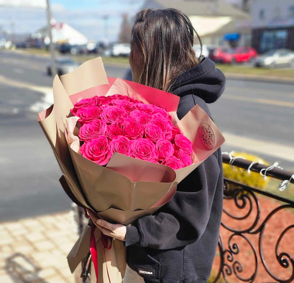 Oversized bouquet of vibrant fuchsia roses wrapped in layered beige and dusty-rose paper with a “Bloom de Fleur” label and tied with matching pink ribbon, held by a person in a dark sweatshirt against a softly blurred suburban street backdrop.