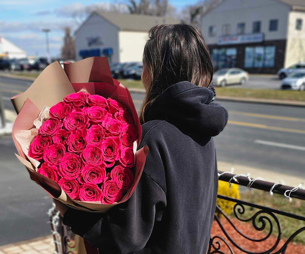 Oversized bouquet of vibrant fuchsia roses wrapped in layered beige and dusty-rose paper with a “Bloom de Fleur” label and tied with matching pink ribbon, held by a person in a dark sweatshirt against a softly blurred  Feasterville-Trevose suburban street backdrop.