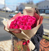 Oversized bouquet of vibrant fuchsia roses wrapped in layered beige and dusty-rose paper with a “Bloom de Fleur” label and tied with matching pink ribbon, held by a person in a dark sweatshirt against a softly blurred suburban street backdrop.