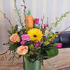 Spring-themed floral arrangement in a square green vase on a wood table, featuring peach garden roses, bright yellow gerbera daisy, hot pink mini carnations and tulips, white larkspur, pussy willow branches, goldenrod and assorted greenery, accented with a yarn‑wrapped carrot ornament.