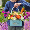 Magenta orchids, purple tulips, peach and orange roses, white lisianthus, pale yellow gerbera daisy and mini carnations, goldenrod sprigs, assorted greenery, and a yarn‑wrapped carrot accent—against a softly blurred outdoor background.
