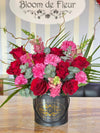 Black Bloom de Fleur hatbox with rope handles holding red roses, pink carnations, snapdragons, eucalyptus leaves, palm fronds.