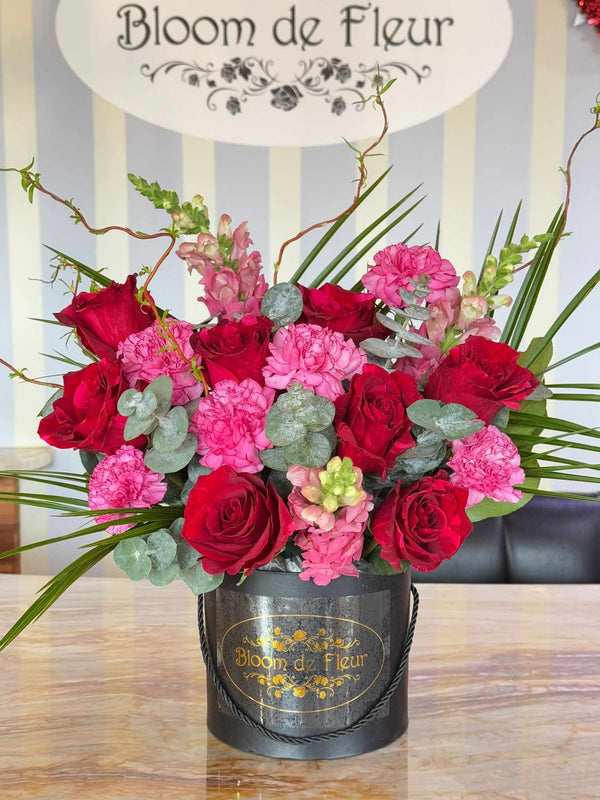 Black Bloom de Fleur hatbox with rope handles holding red roses, pink carnations, snapdragons, eucalyptus leaves, palm fronds.