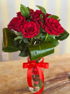 Clear glass vase with red heart pattern and tied red ribbon, holding six deep red roses and folded green leaves.