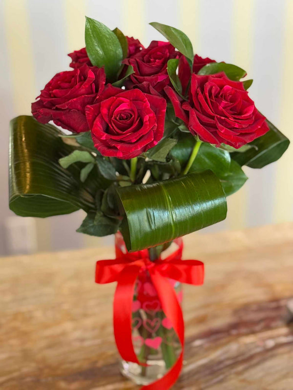 Clear glass vase with red heart pattern and tied red ribbon, holding six deep red roses and folded green leaves.