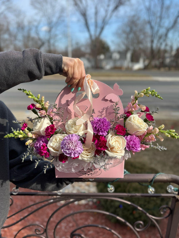 Pink purse-style Bloom de Fleur box with handle, filled with ivory roses, purple and fuchsia carnations, plus pink snapdragons
