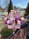 Pink purse-style Bloom de Fleur box with handle, filled with ivory roses, purple and fuchsia carnations, plus pink snapdragons