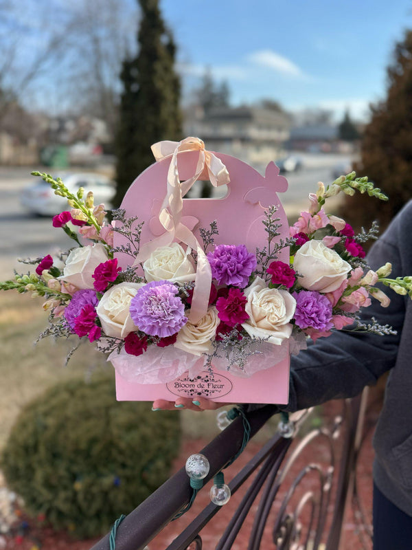Pink purse-style Bloom de Fleur box with handle, filled with ivory roses, purple and fuchsia carnations, plus pink snapdragons