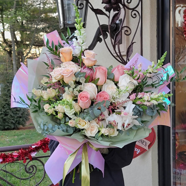 Luxury pastel mixed-flower bouquet featuring soft pink, peach, and ivory roses, white cymbidium orchids, snapdragon spikes, Queen Anne’s lace, astilbe sprigs, and silver dollar eucalyptus greenery, all artfully arranged and wrapped in iridescent and sage-green tissue with a gold ribbon bow—ideal for weddings, anniversaries, or special occasions.