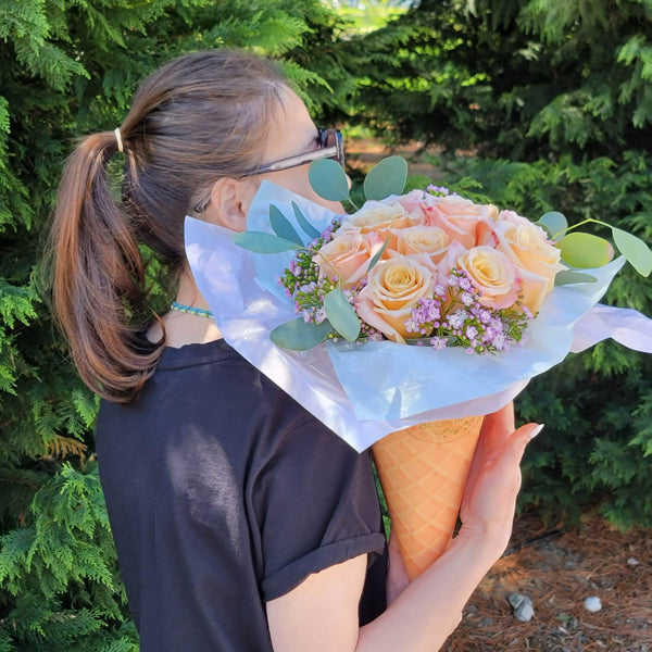 A person in a black knotted T‑shirt holds a waffle‑cone‑shaped Bloom de Fleur vessel wrapped in white and pale blue tissue paper, filled with peach‑cream roses, clusters of tiny lavender waxflowers, and eucalyptus sprigs, set against a backdrop of evergreen trees and pine‑straw ground.