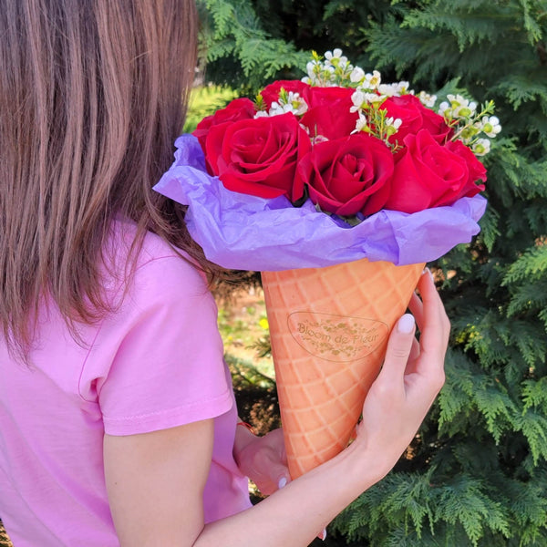 A person in a pink T‑shirt holds a waffle‑cone‑shaped Bloom de Fleur vessel wrapped in purple tissue, filled with a bouquet of deep red roses and sprigs of white waxflower, set against a backdrop of dense evergreen foliage.