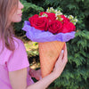 A person in a pink T‑shirt holds a waffle‑cone‑shaped Bloom de Fleur vessel wrapped in purple tissue, filled with a bouquet of deep red roses and sprigs of white waxflower, set against a backdrop of dense evergreen foliage.