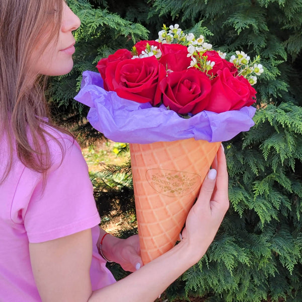 A person in a pink T‑shirt holds a waffle‑cone‑shaped Bloom de Fleur vessel wrapped in purple tissue, filled with a bouquet of deep red roses and sprigs of white waxflower, set against a backdrop of dense evergreen foliage.