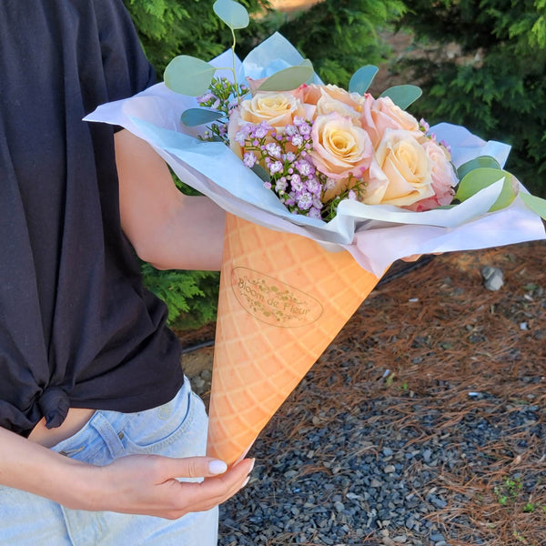 A person in a black knotted T‑shirt holds a waffle‑cone‑shaped Bloom de Fleur vessel wrapped in white and pale blue tissue paper, filled with peach‑cream roses, clusters of tiny lavender waxflowers, and eucalyptus sprigs, set against a backdrop of evergreen trees and pine‑straw ground.