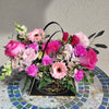 A low, lush arrangement in a black triangular “Bloom de Fleur” gift box with twin black rope handles, sitting on a blue‑green mosaic tile table against a pale stucco wall. The box is filled with hot‑pink roses, pale pink carnations, soft pink gerbera daisies, bright fuchsia spray carnations, clusters of tiny white waxflowers, and white snapdragon blossoms, all accented with fern fronds and eucalyptus leaves.