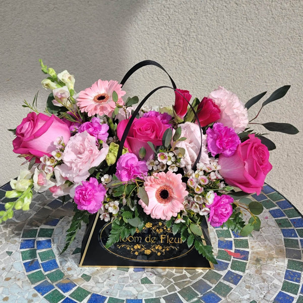 A low, lush arrangement in a black triangular “Bloom de Fleur” gift box with twin black rope handles, sitting on a blue‑green mosaic tile table against a pale stucco wall. The box is filled with hot‑pink roses, pale pink carnations, soft pink gerbera daisies, bright fuchsia spray carnations, clusters of tiny white waxflowers, and white snapdragon blossoms, all accented with fern fronds and eucalyptus leaves.