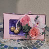 A blush‑pink rectangular “Bloom de Fleur” gift box with rose‑gold metallic trim sits on a blue‑green mosaic tile table against a stucco wall; through its left window you see three gold‑foil wrapped chocolates nestled in purple tissue paper, while the right side is styled with two coral‑pink roses, a white orchid blossom, sprigs of eucalyptus, and a pale pink ribbon tied in a bow.