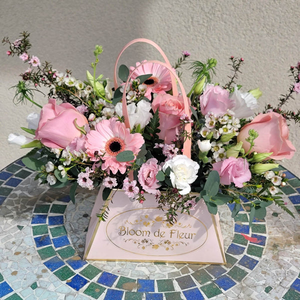 A pale pink triangular “Bloom de Fleur” gift box with gold foil logo and twin pink ribbon handles sits on a blue‑green mosaic tile table against a light stucco wall. It’s overflowing with a lush mix of blush and hot‑pink roses, pale pink gerbera daisies, white carnations, tiny pink waxflower sprigs, soft snapdragon buds, and accents of fern fronds and eucalyptus leaves.
