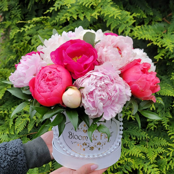 A pair of hands holding a white round “Bloom de Fleur” hatbox—adorned with a string of faux pearls and a gold scrollwork logo—overflowing with lush hot‑pink, coral, and pale blush peonies nestled in glossy green leaves, set against a backdrop of dense evergreen foliage.